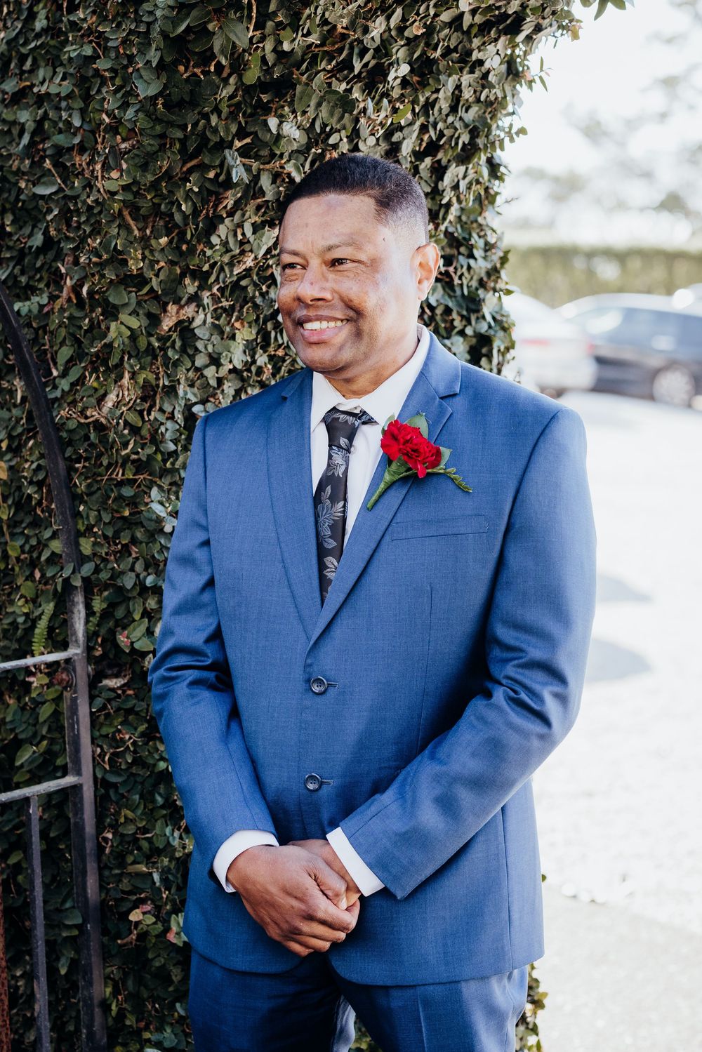 Person in a blue suit with a red boutonniere smiling during an outdoor wedding event.