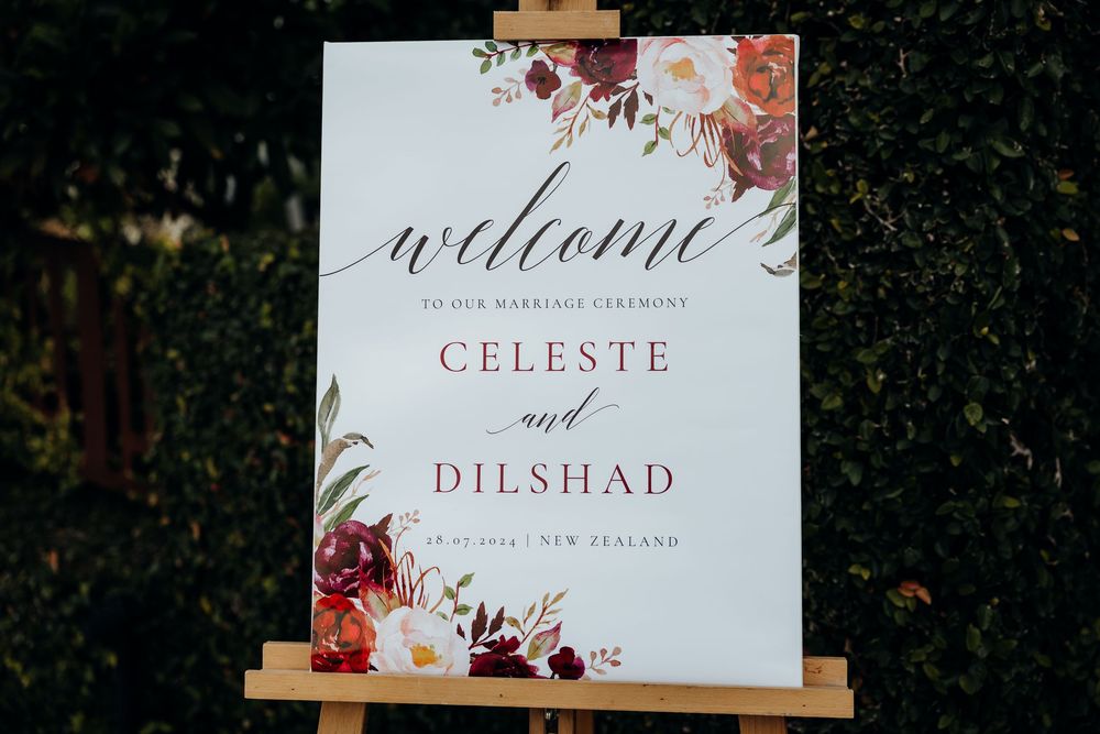 Elegant wedding welcome sign with floral design and calligraphy displayed on wooden easel against green foliage background.