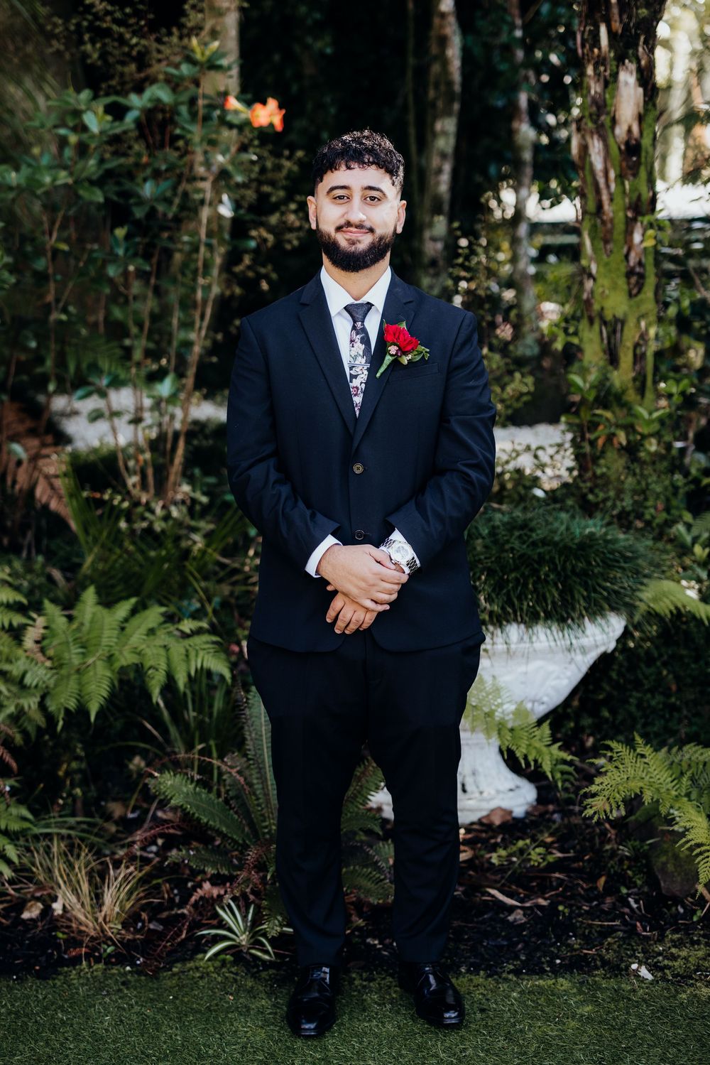 A person in a dark suit with a red boutonniere sits on a bench in a garden setting.