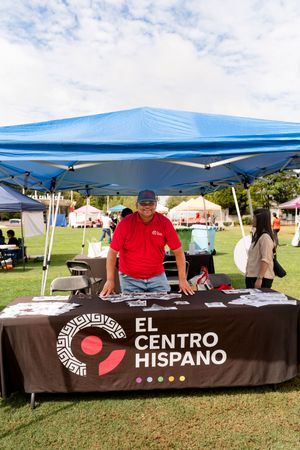 Outdoor vendor booths with brown tablecloths and blue tents displaying El Centro Hispano branding at a community event.