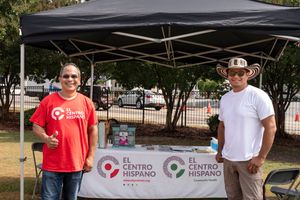 Two people stand behind a table with a banner at an outdoor event under a black pop-up tent.