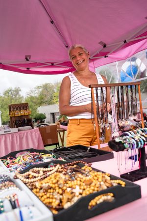 Vendor displays colorful jewelry and accessories under a pink tent at an outdoor market.