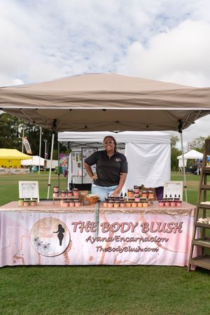 Pink-themed vendor booth displaying The Body Blush branding and products under white canopy tents on grass.