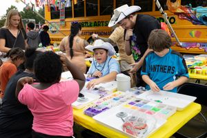 Children and adults participate in arts and crafts activities at yellow tables near school bus.