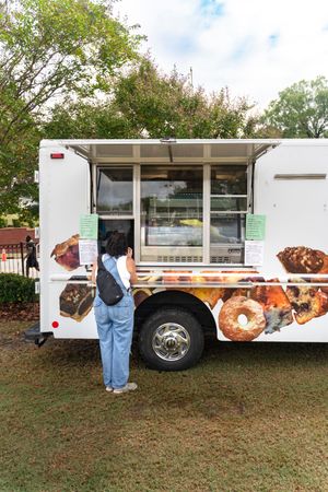 A white food truck decorated with donut graphics parked on grass with a customer at the window.