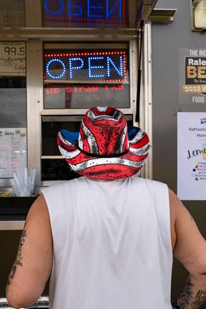 Person wearing a white tank top and patriotic red white and blue cowboy hat stands in front of an open sign.