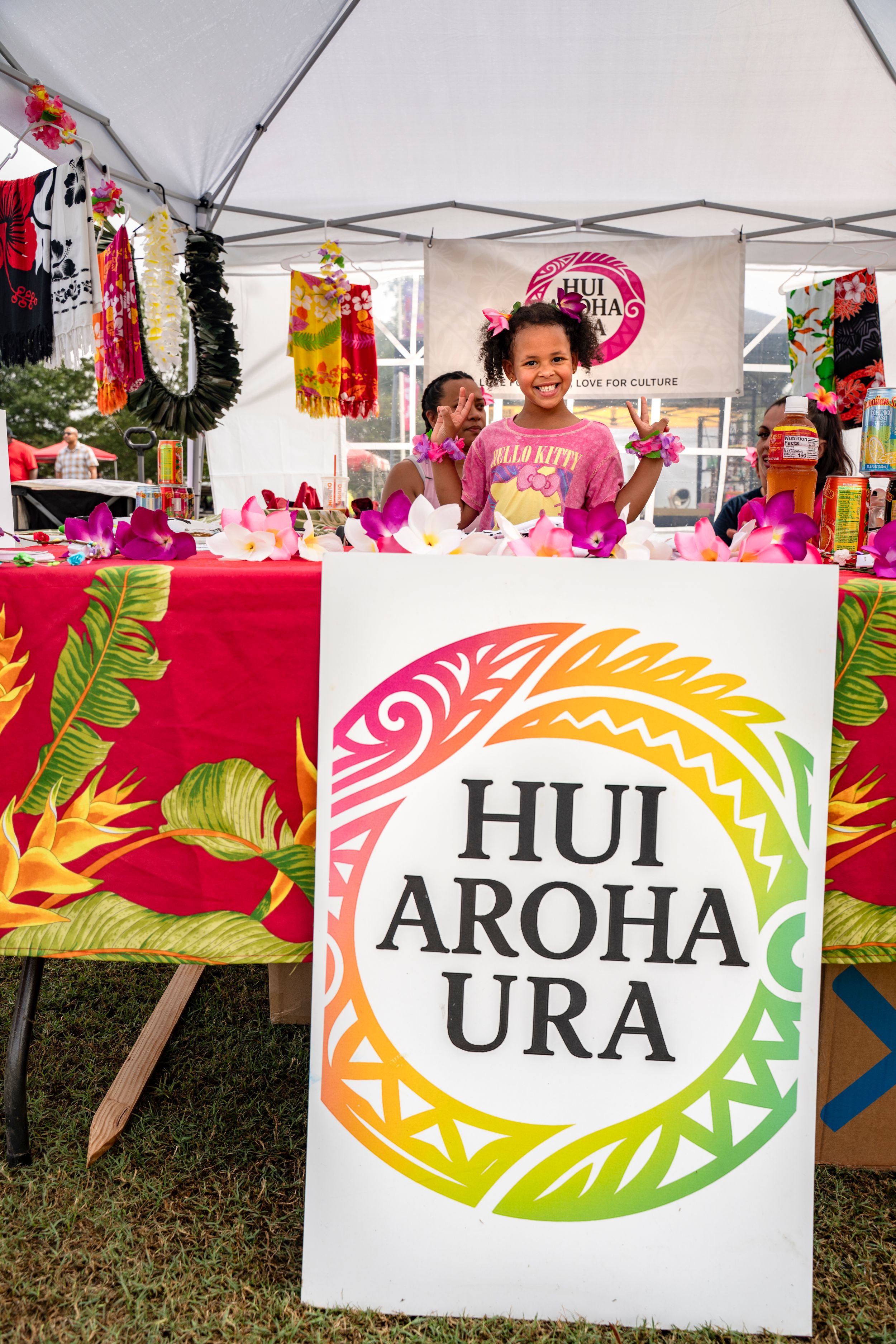 Colorful vendor booth banner displaying 'Hui Aroha Ura' with tropical leaf designs at an outdoor festival.