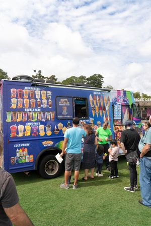 Customers line up at colorful food trucks serving frozen treats on a grassy field under cloudy skies.
