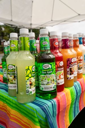 Colorful array of bottled drinks displayed on rainbow-striped tablecloth at outdoor event.