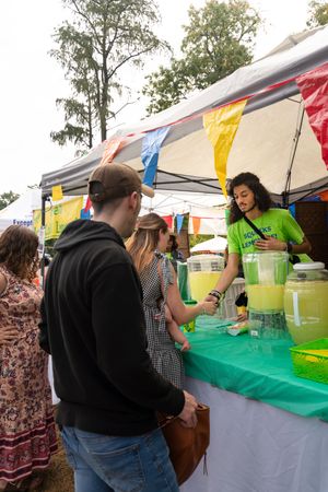 People gathered around a green-covered table serving drinks at an outdoor festival.