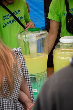 Row of lemonade dispensers with bright yellow drinks being served by volunteers in green shirts.
