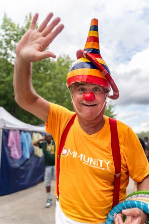 A person in an orange community t-shirt wearing a striped hat and red clown nose waves at the camera.