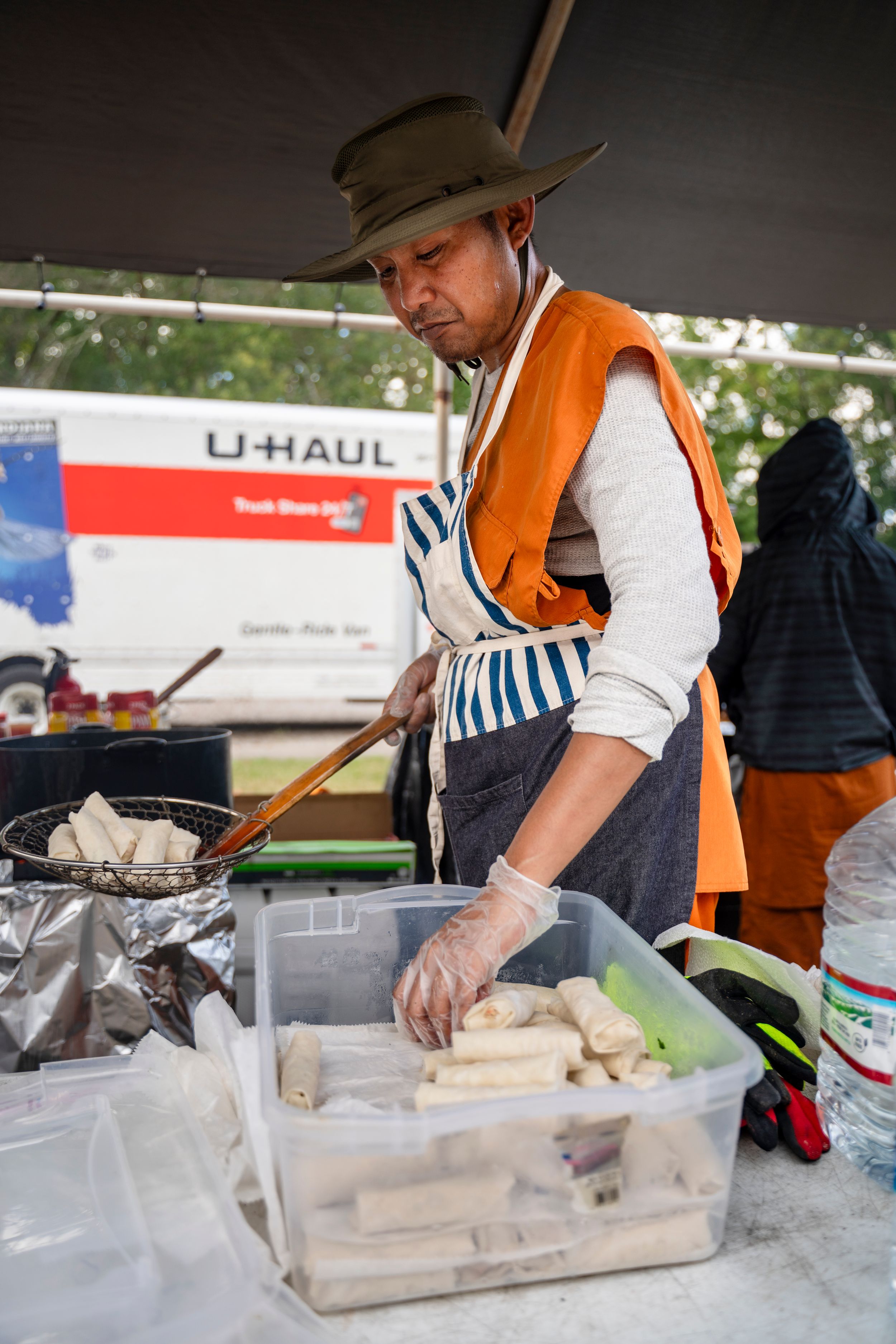 Vendor in orange shirt and hat prepares food at an outdoor event station.