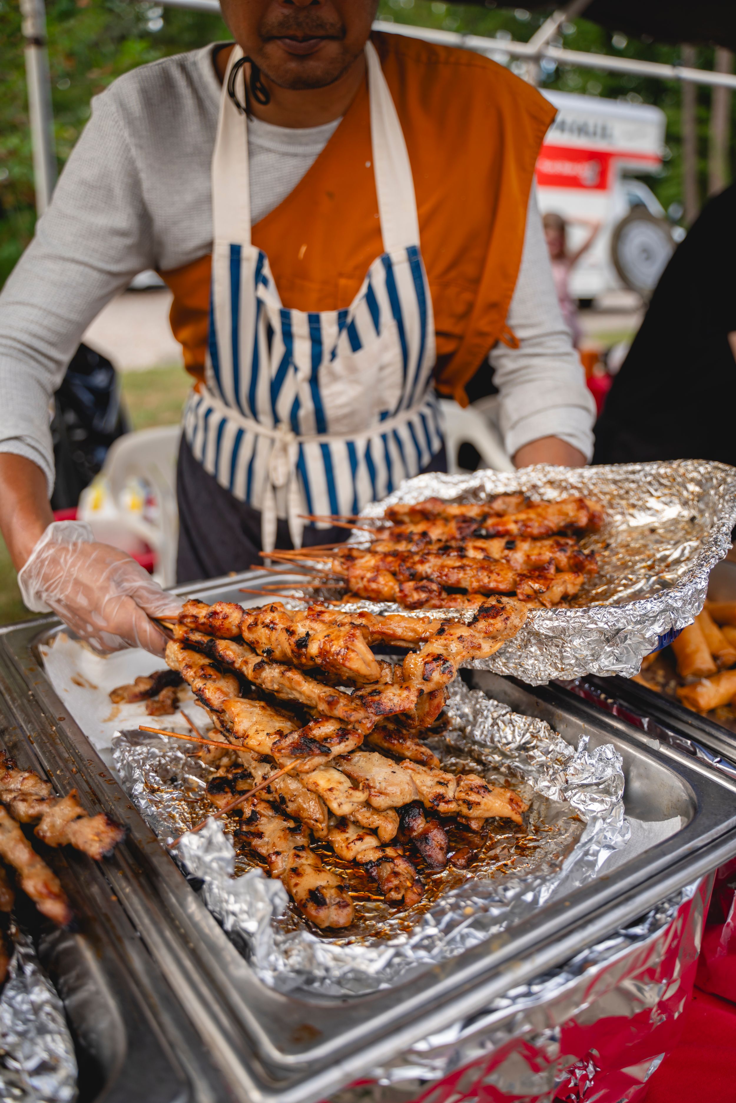 Grilled meat on foil-lined trays being served at an outdoor food festival.