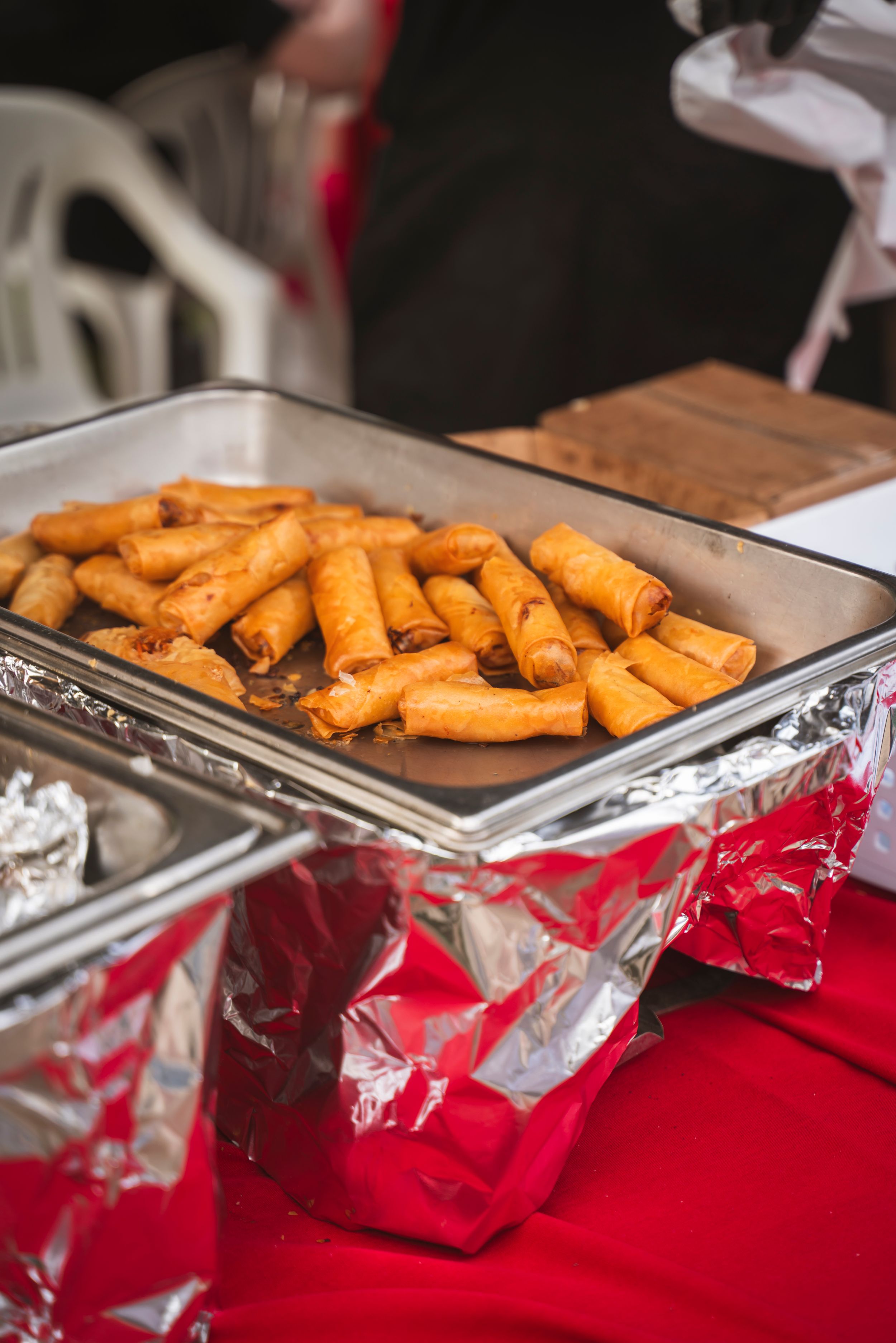 Freshly cooked spring rolls arranged in rows on metal serving trays with red tablecloth underneath.