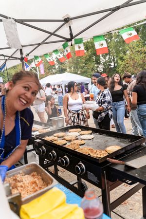 People grilling food on large outdoor griddles under a tent with festive flags.