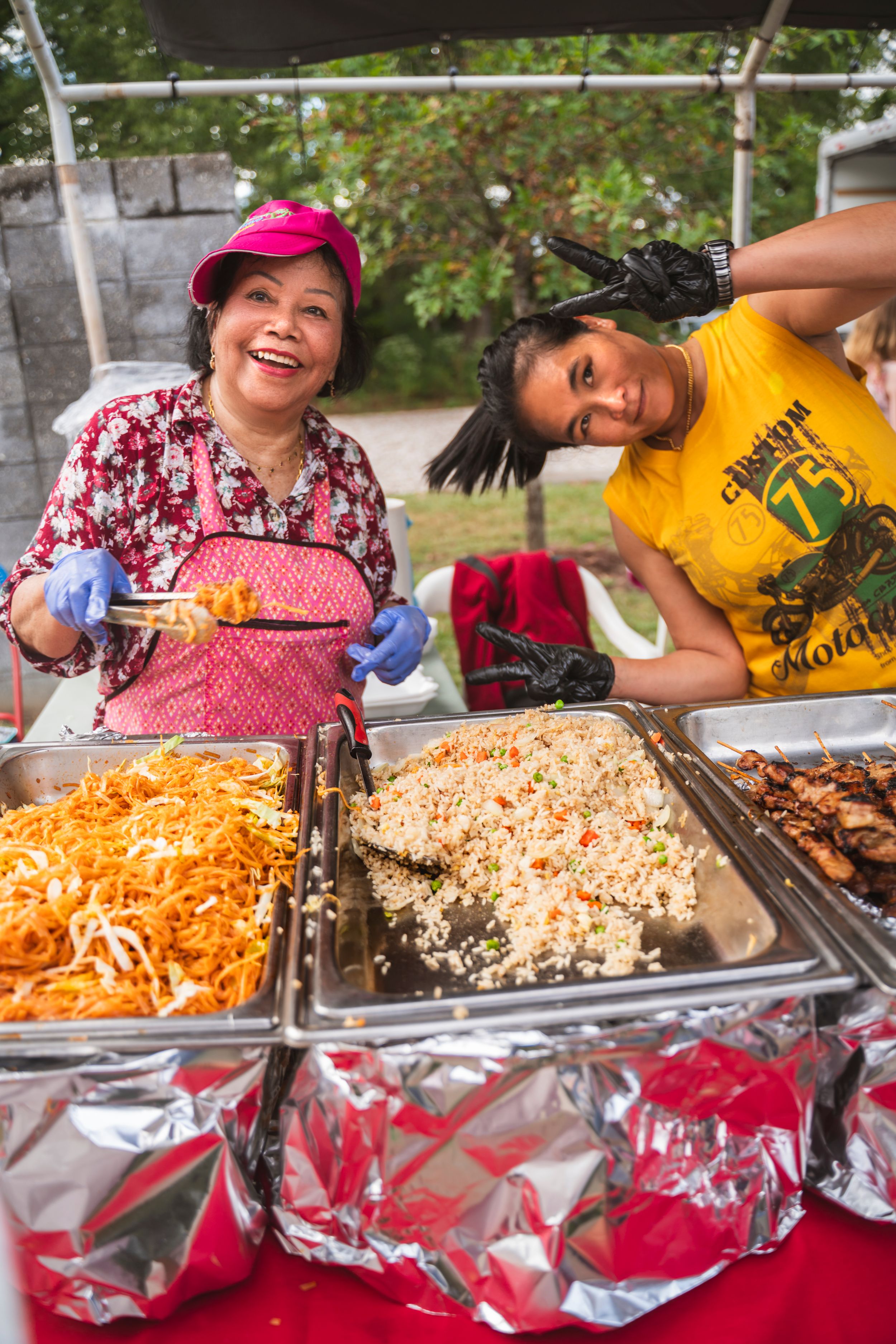 Food servers dish out colorful noodles and rice from metal catering trays at an outdoor event.