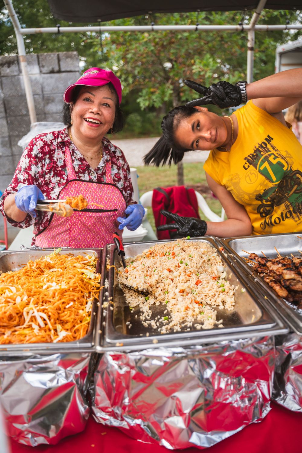 Food servers dish out colorful noodles and rice from metal catering trays at an outdoor event.