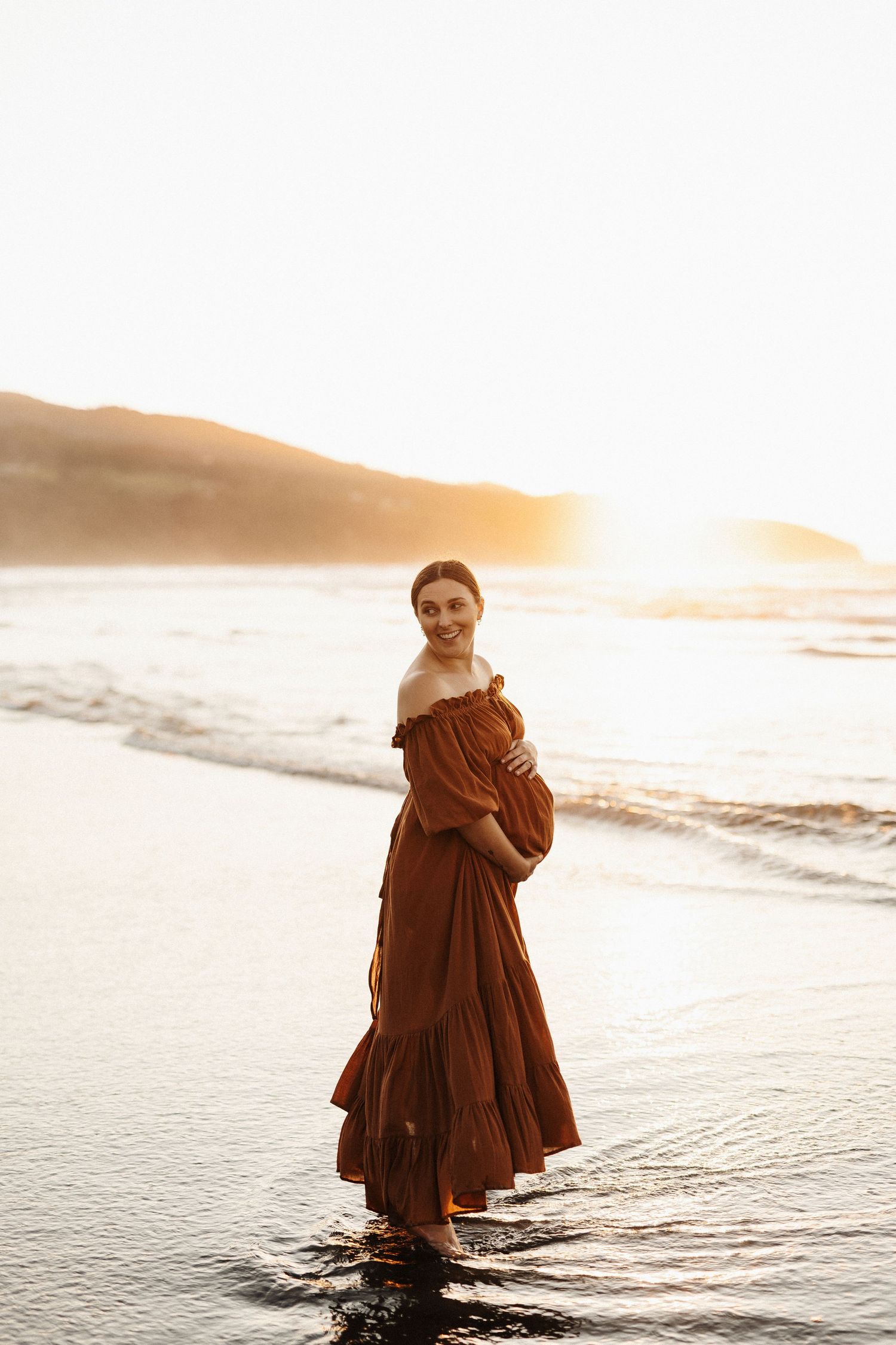 A person in a brown maxi dress stands in shallow ocean water at sunset.
