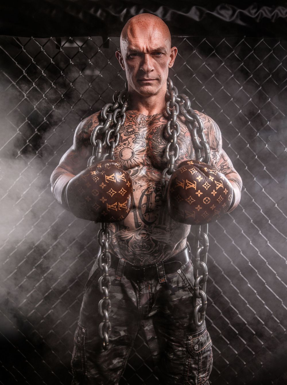 Muscular fighter with chains and boxing gloves poses against chain-link fence in dramatic lighting.