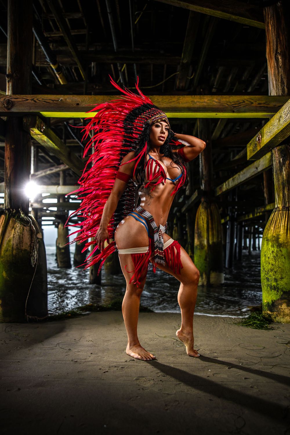 A striking dance performance under a pier featuring a dramatic red feathered headdress and vibrant costume styling.