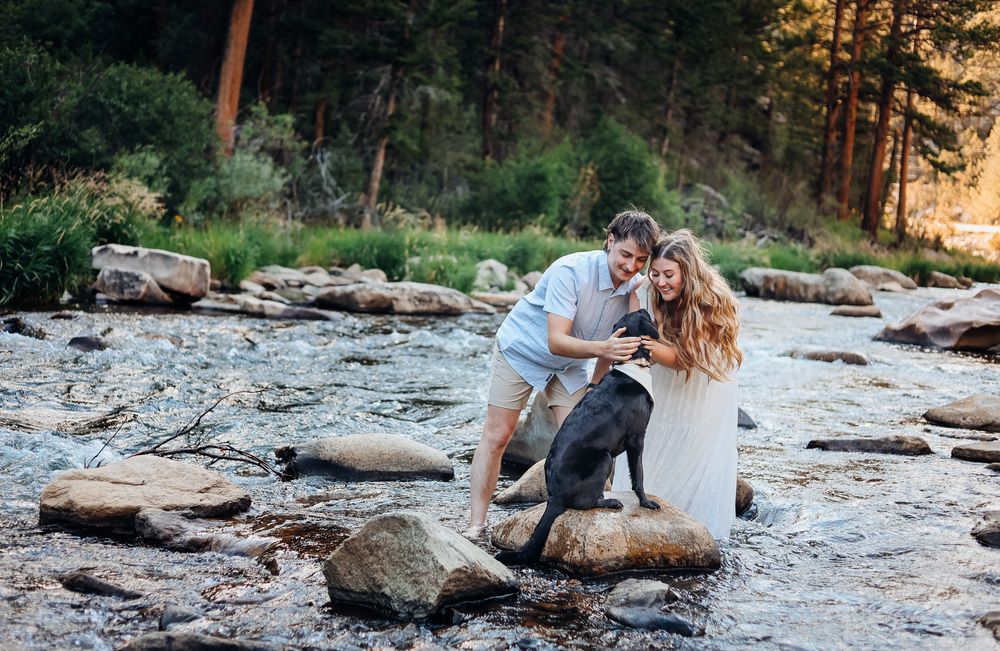 Couple and black dog playing on rocky river with lush forest backdrop in a romantic outdoor setting.