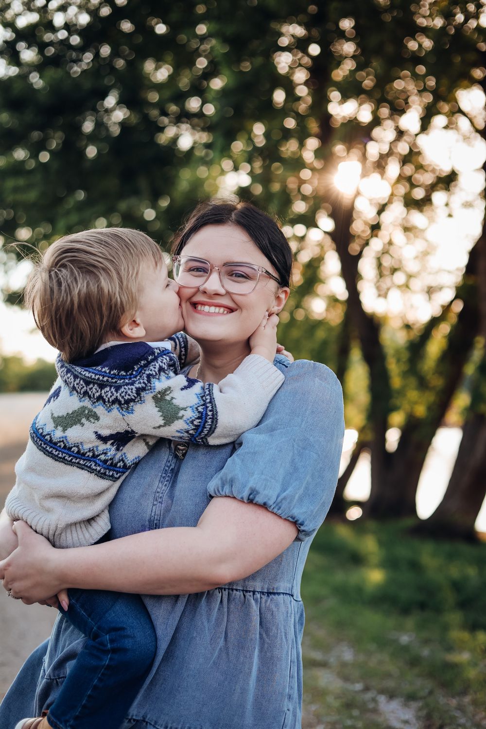 A smiling woman in glasses holds a young boy who kisses her cheek outdoors in golden sunlight near trees.