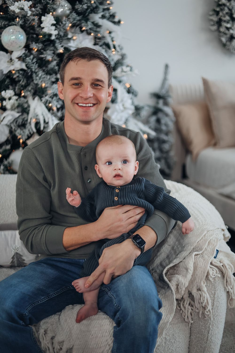 Heartwarming indoor holiday photoshoot featuring an infant in a navy outfit on a beige couch with decorated Christmas trees. Family studio