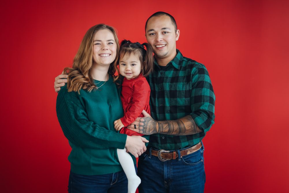 Holiday family portrait session against vibrant red backdrop with coordinated green and plaid outfits. Family studio photography