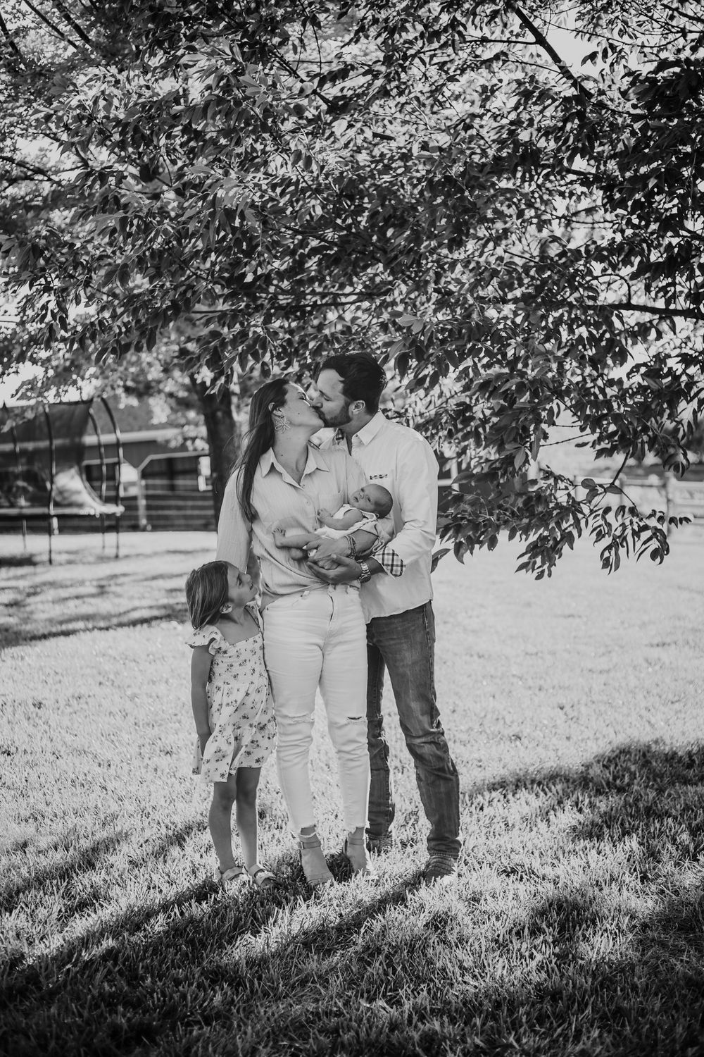 Black and white family photo of couple kissing while holding newborn baby, with young daughter standing beside them outdoors.