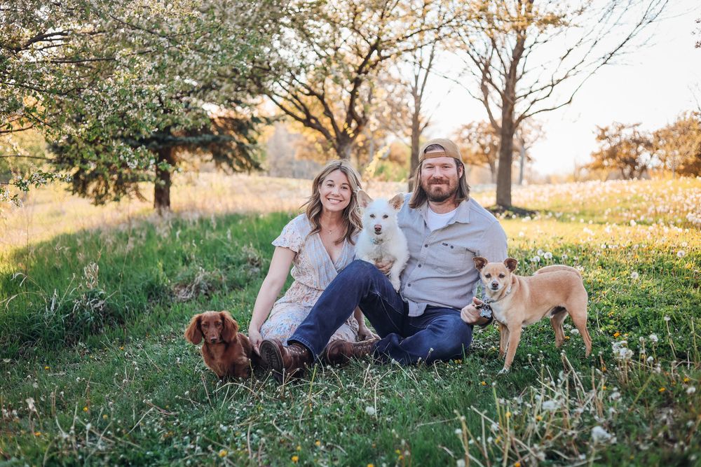 Couple sitting on grass with three dogs, including a dachshund and white dog, in a sunny outdoor park setting.