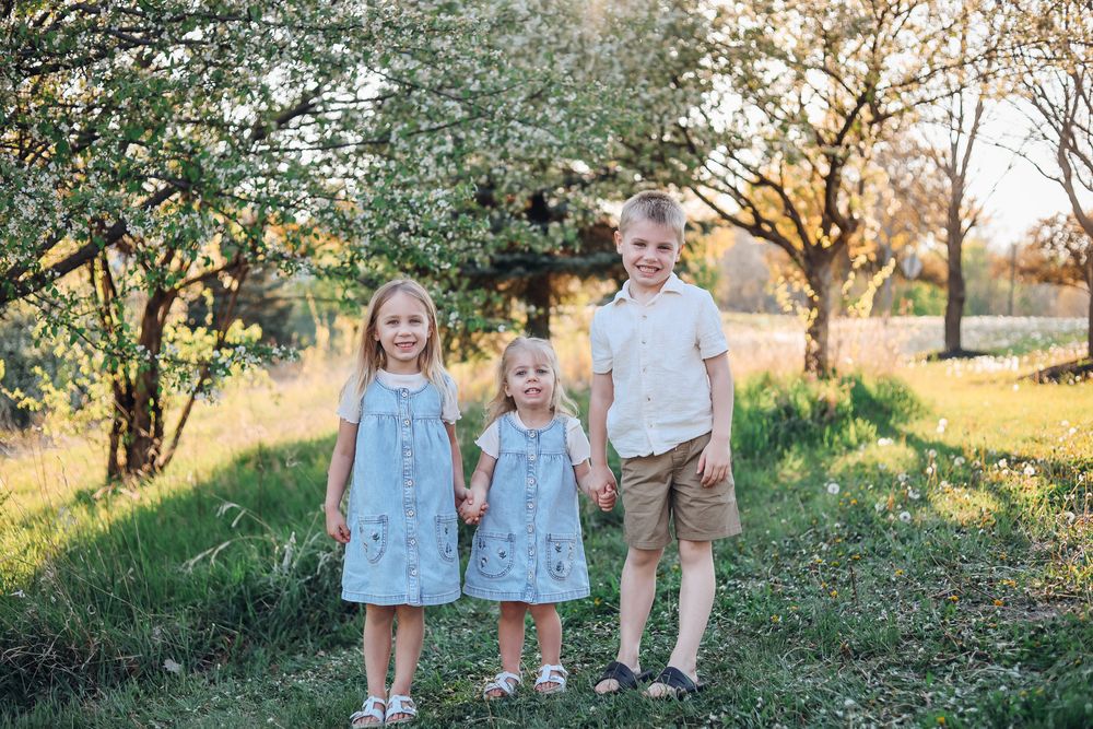 Three young children holding hands in a sunny garden with blooming trees, two girls in blue dresses and a boy in khaki shorts.