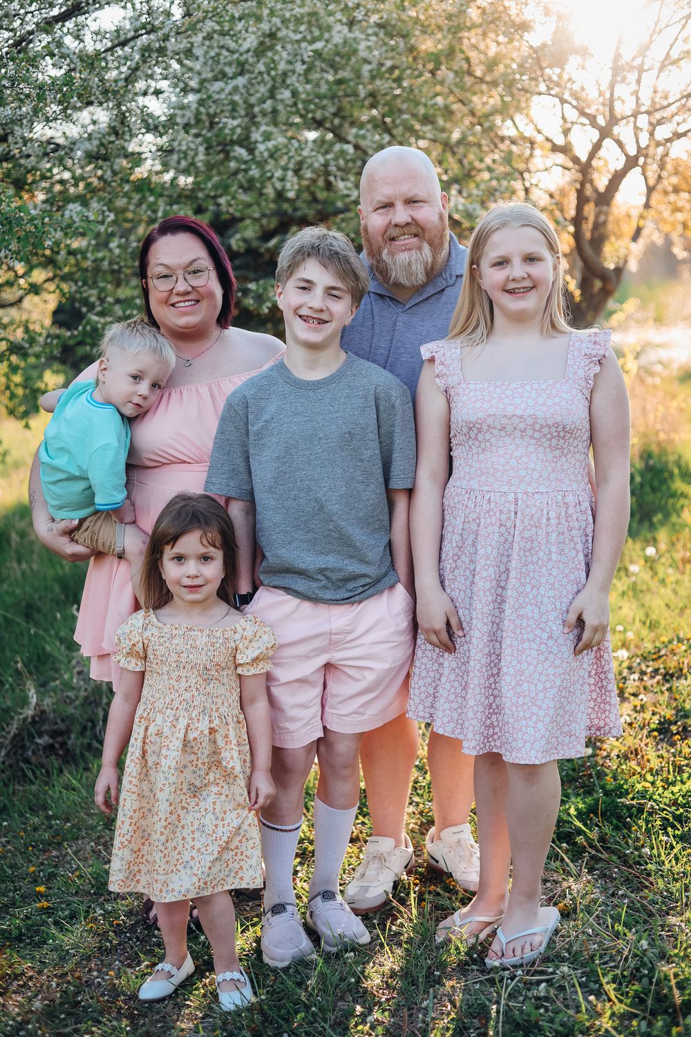 Family of six posing outdoors near blooming trees at golden hour, parents with four children dressed in pink and pastel tones.