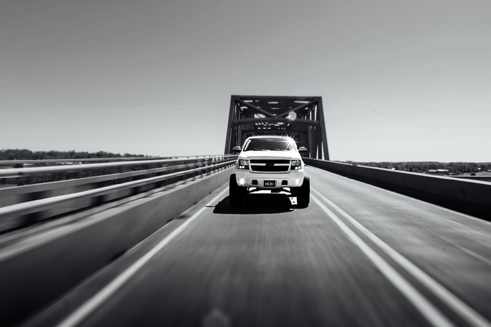 Black and white photo of SUV crossing bridge with motion blur effect.