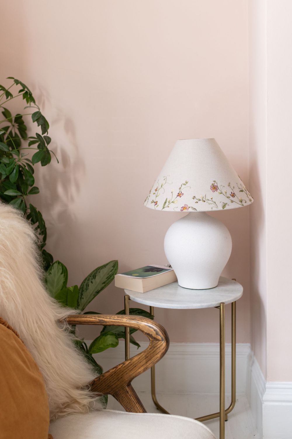 White ceramic table lamp with floral pattern shade sits atop gold metal side table against blush pink wall with greenery.
