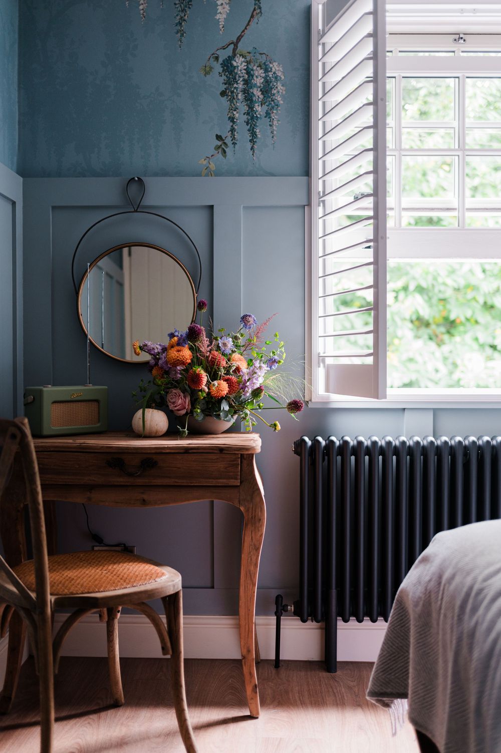A rustic wooden console table with floral arrangement sits against a blue-gray wall with vintage radiator and bright windows.