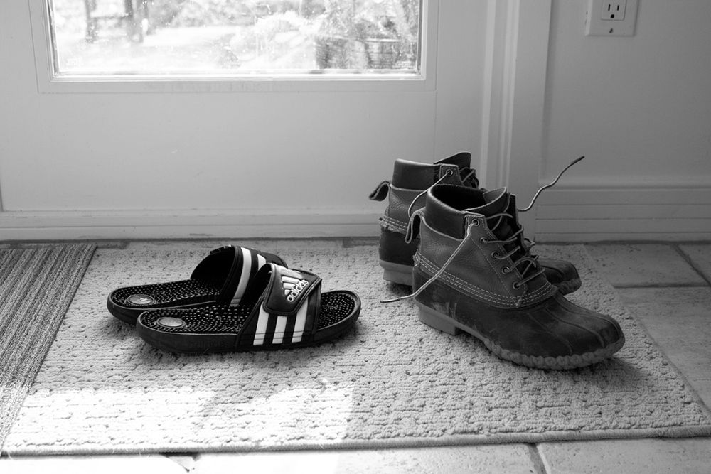 Black and white photo of shoes on a doormat by a window showing Adidas slides and leather boots.