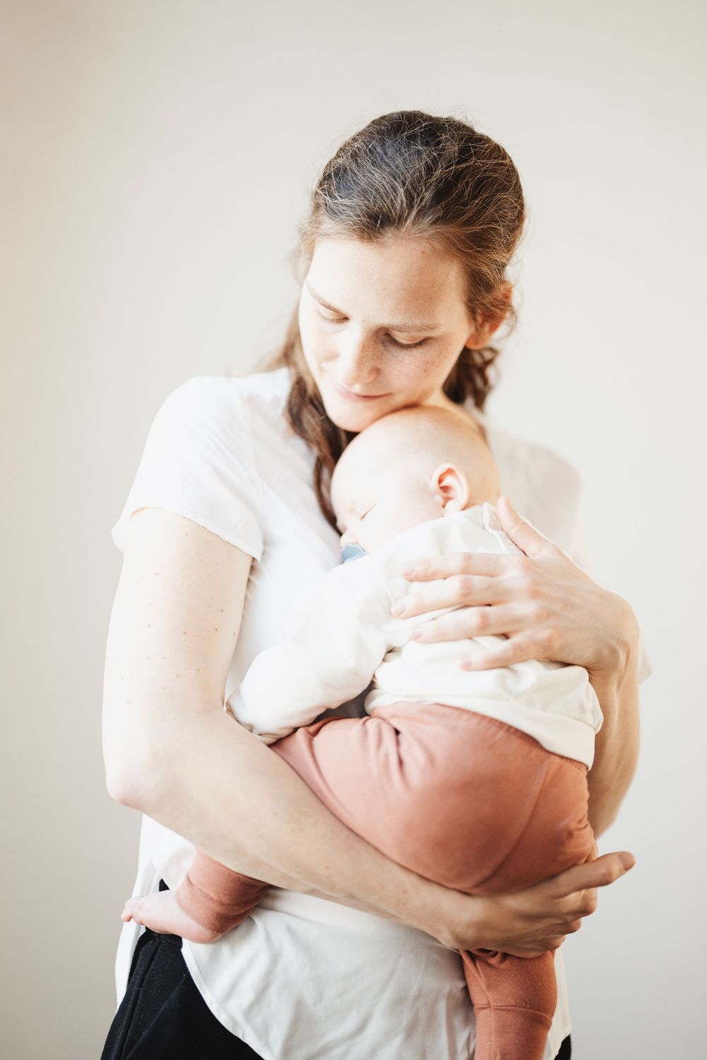Mother holds sleeping baby dressed in white top and pink pants against white wall backdrop.
