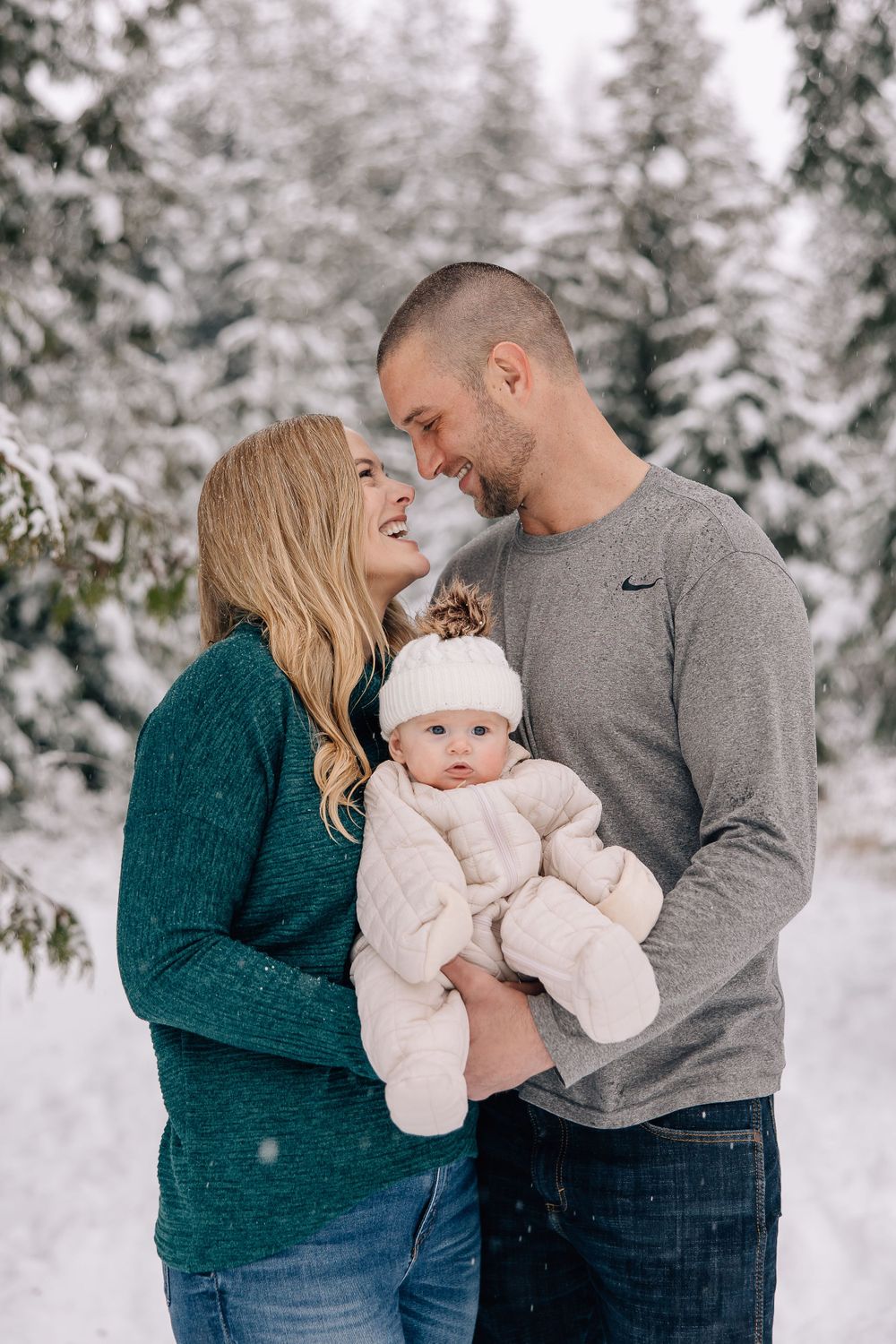 A couple shares a joyful moment with their baby in a snowy winter forest setting, wearing cozy sweaters.