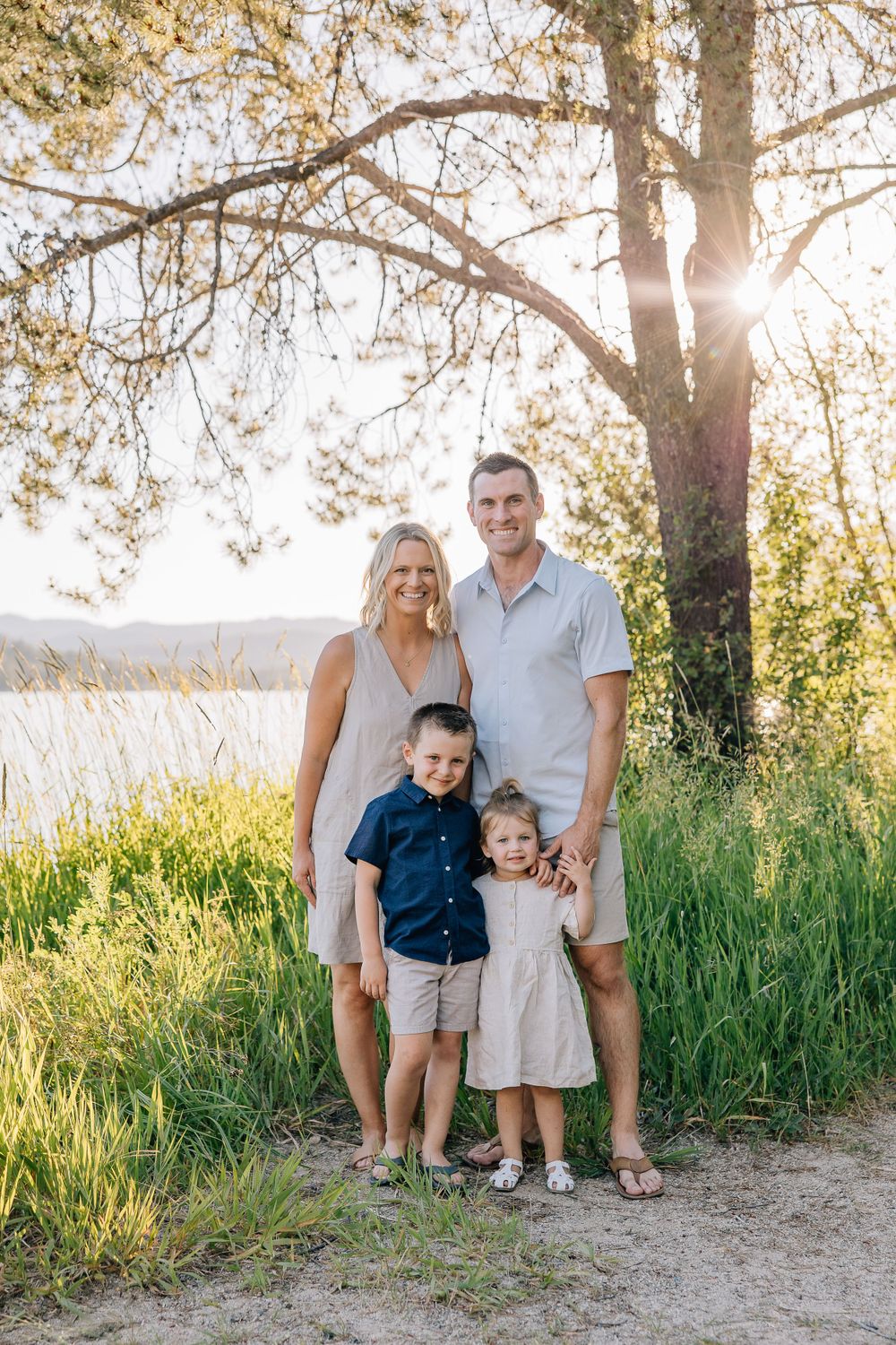 Family portrait in summer setting with sunlight streaming through tree branches near lake.