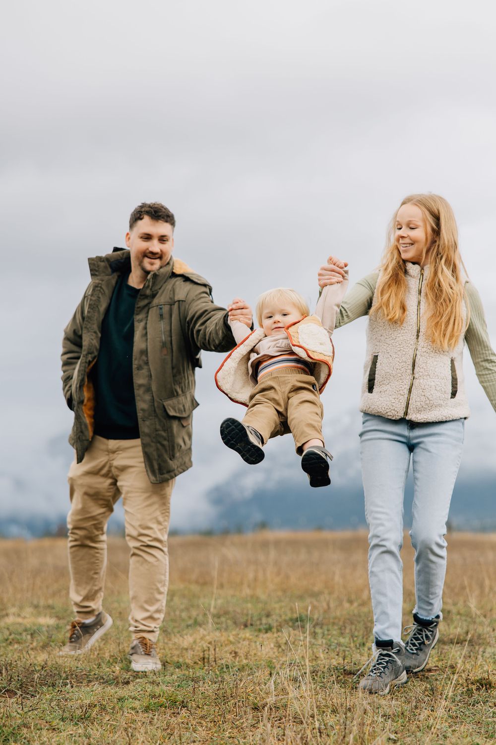 Parents playfully swing child while walking in field under cloudy sky.