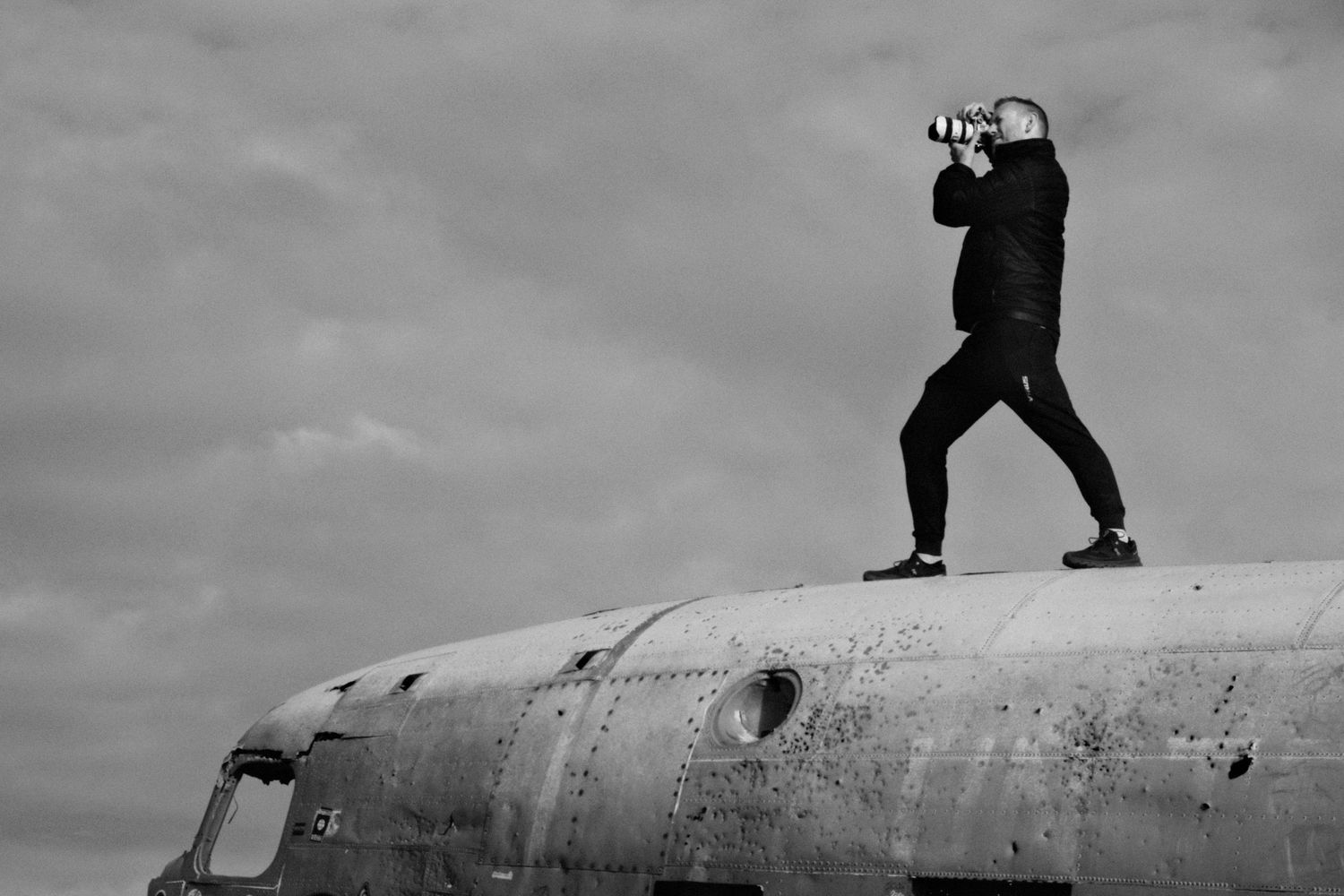 Silhouetted photographer stands atop weathered aircraft fuselage against dramatic cloudy sky in black and white.
