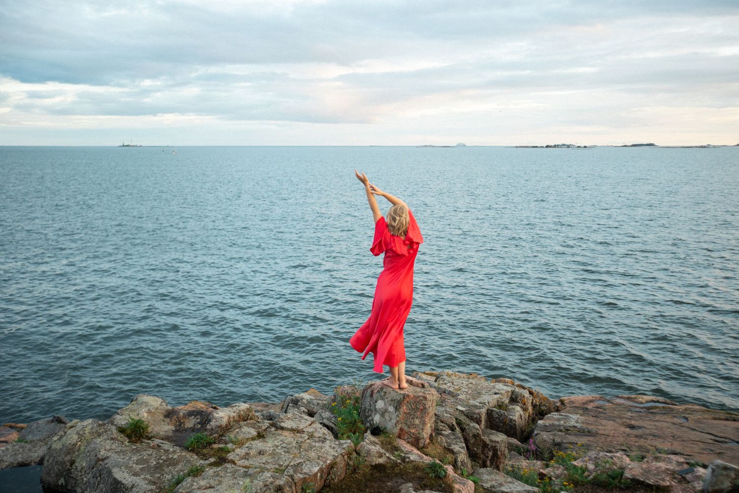 A person in a flowing red dress stands gracefully on rocky coastal cliffs overlooking a serene ocean view.