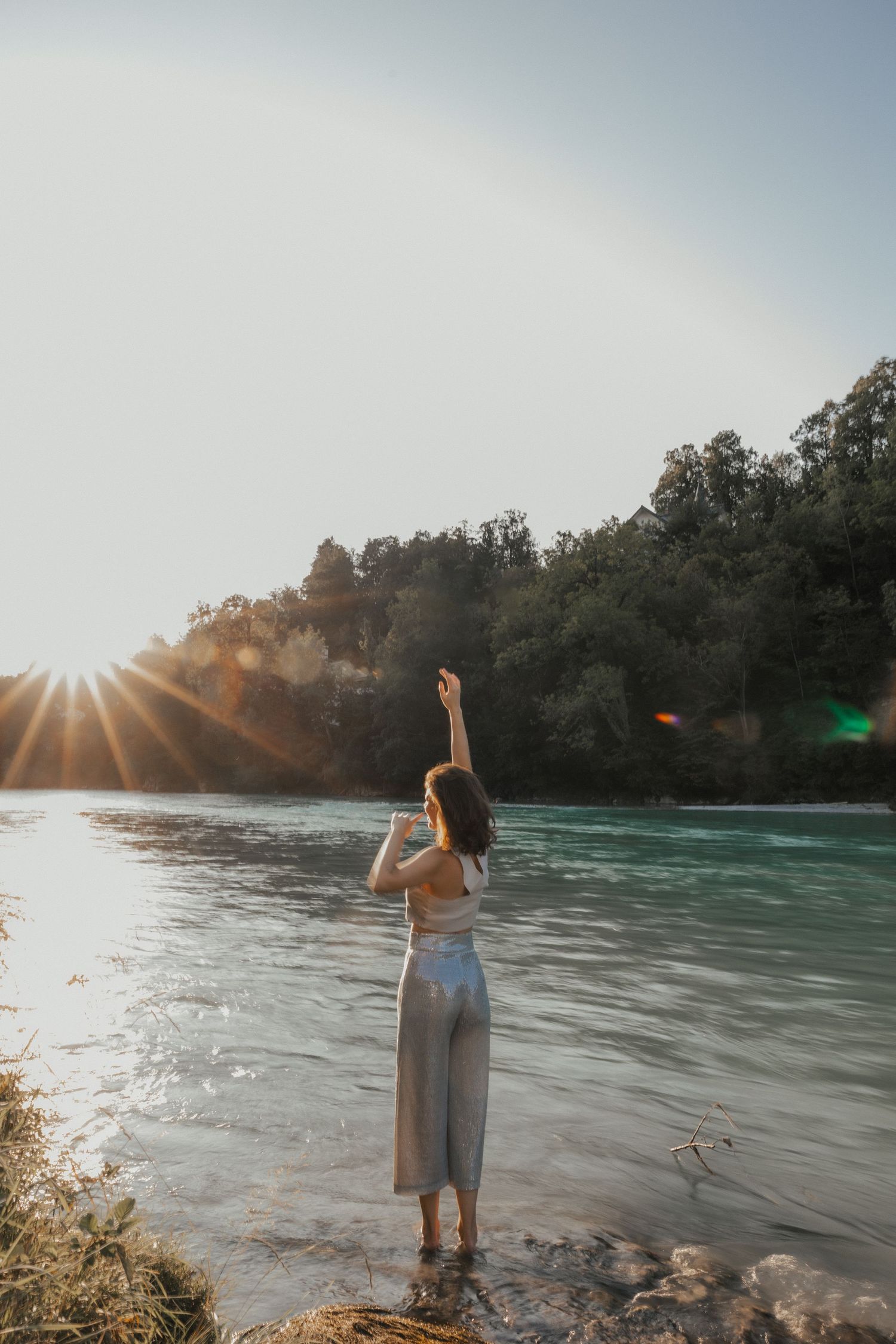 Person standing by riverbank during golden hour with sunburst through trees.
