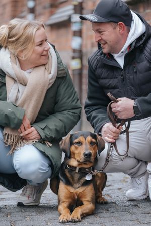 Mensch-Hund-Fotografie: Paar hockt mit seinem Hund auf einem Gehweg im urbanen Umfeld von München.