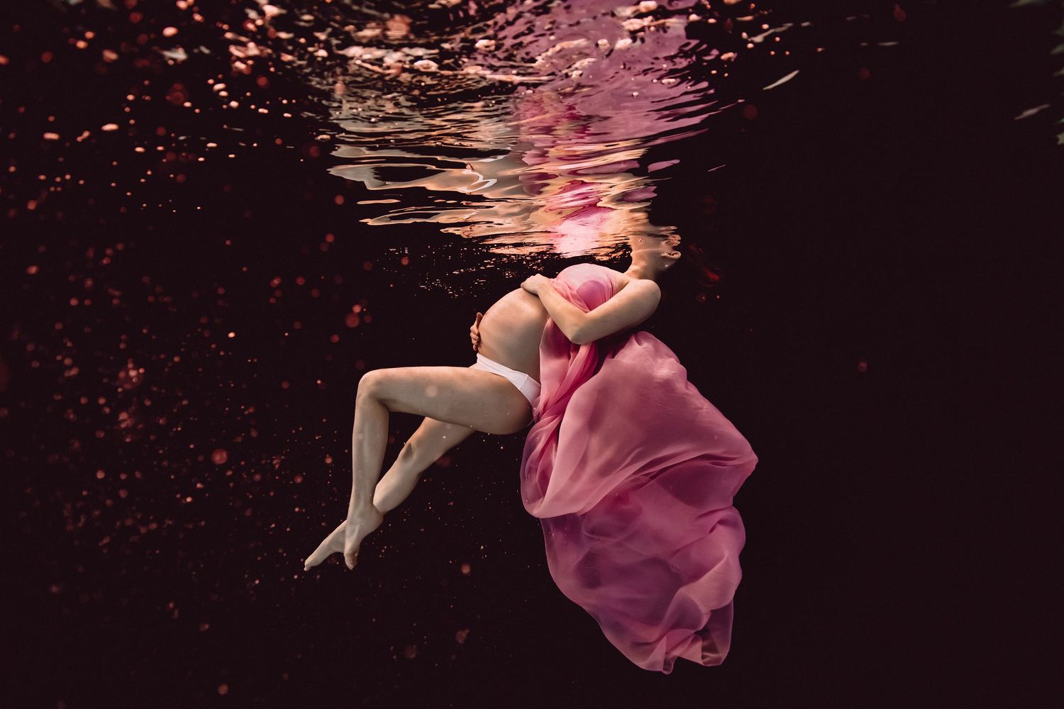 Pregnant woman in pink flowing dress floating gracefully underwater with pink particles in dark water.