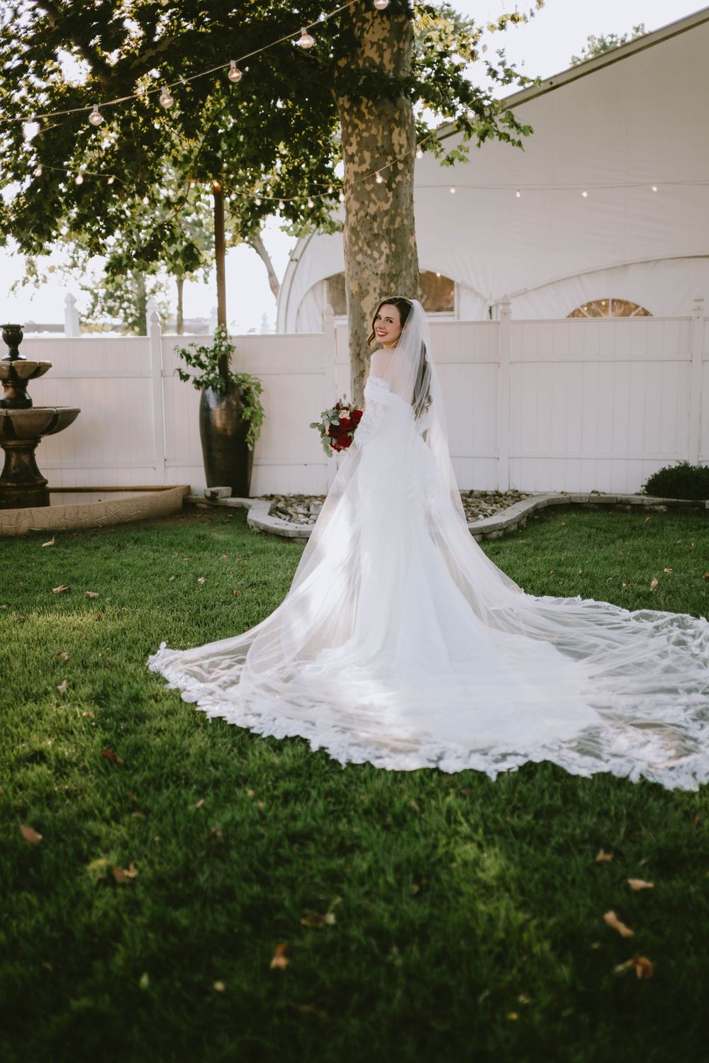 Bride in a flowing white lace wedding gown with cathedral veil holding red bouquet, posing in lush garden.