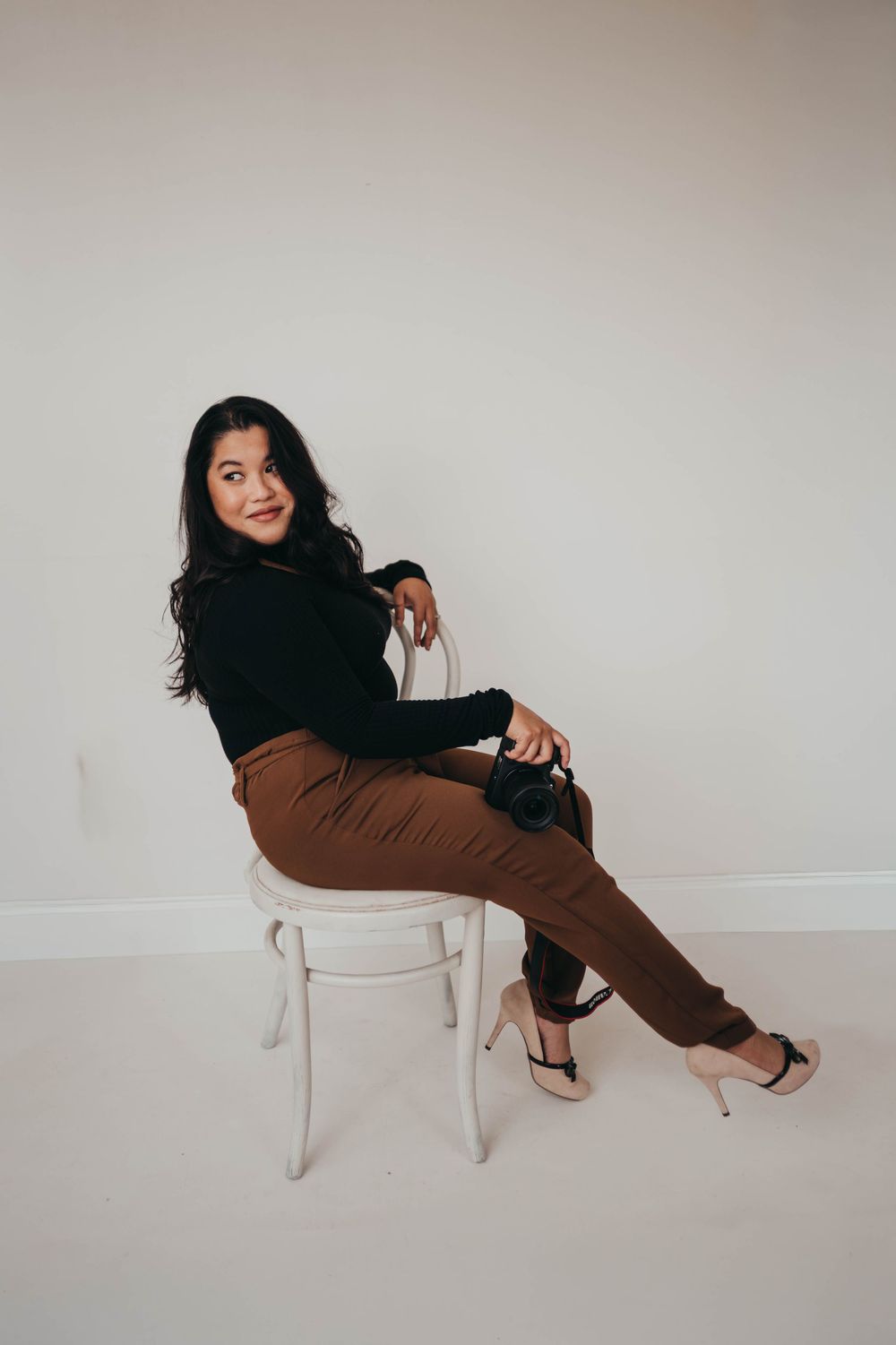Woman in black top and brown pants sits on white chair holding camera, smiling over shoulder.