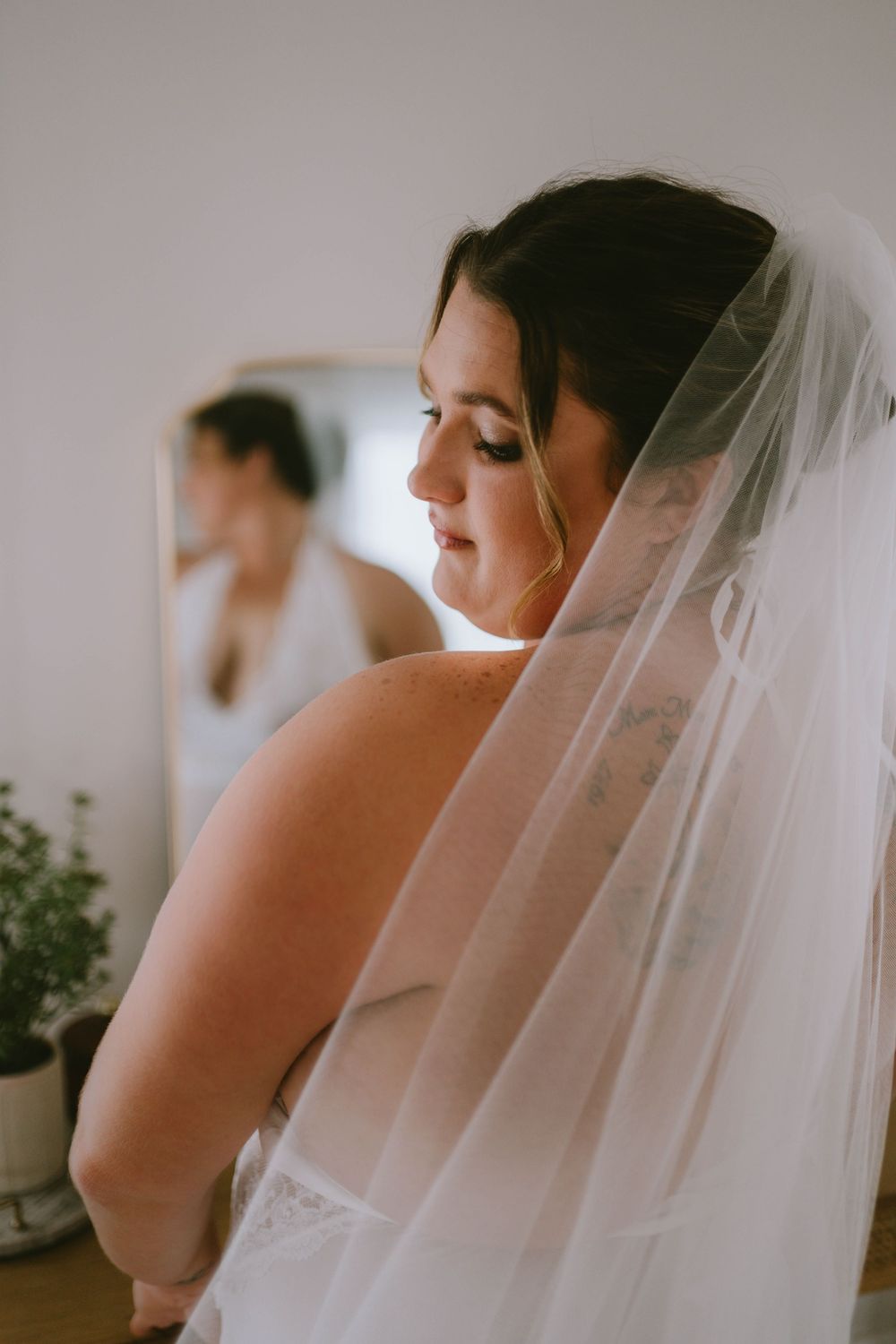 Bride in white wedding dress and veil gazes down, with mirror reflection visible in soft natural light.