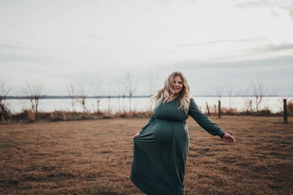 Pregnant woman in green dress twirling in a field near a lake on an overcast day.